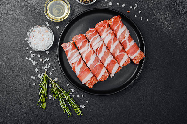 istock Raw minced meat rolls wrapped in bacon on black plate with spices 2233325366