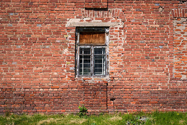 Old brick wall with a weathered window and grass at the base. The surface features aged textures, cracks, and faded mortar