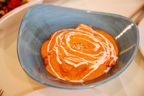 Elevated view of metal bowl of Indian butter chicken curry sauce with cream drizzled on the surface, lachha paratha, multi layered Indian flat bread and a bowl of boiled white rice on a wooden table.