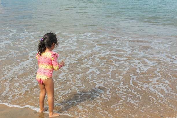 A carefree little girl stands on the sandy beach, her hair tousled by the ocean breeze. Sun-kissed and joyful, she gazes out at the horizon.