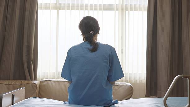 istock Sad patient and thinking stress anxiety with symptoms of depression and sadness, problems, mentally ill person. Woman in a blue patient uniform gown sits alone on a bed in a private room and looking out the window in hospital, expressing her concern about 2233342619