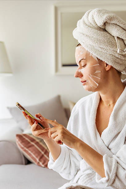 istock Relaxed Woman Applying Face Mask While Browsing Smartphone at Home 2233350973