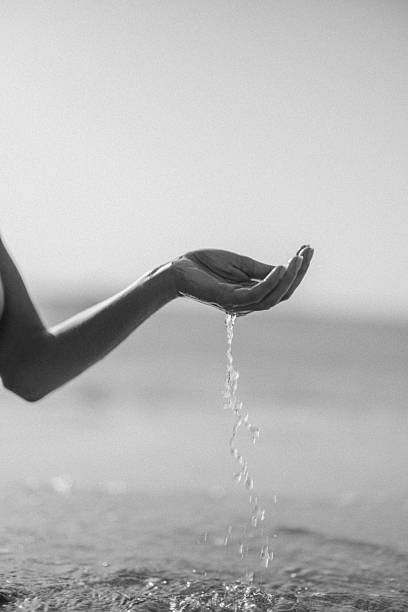 A monochrome image capturing a human hand with water streaming from it by the beach. The simplicity of the black and white composition emphasizes emotion, moments, and reflection related to nature, tranquility, and purity.