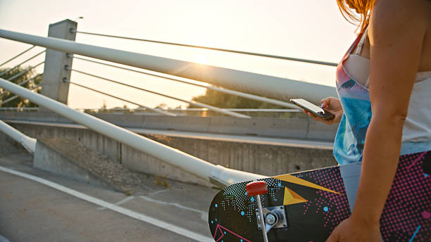 istock Young Woman Using Smartphone while Holding Skateboard Under Modern Bridge at Sunset 2233363883