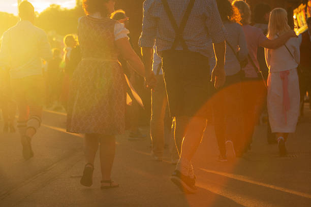 A warm sunset illuminates a couple holding hands, dressed in traditional Bavarian costumes during Oktoberfest, surrounded by a lively festive crowd enjoying the celebration's joyous atmosphere.
