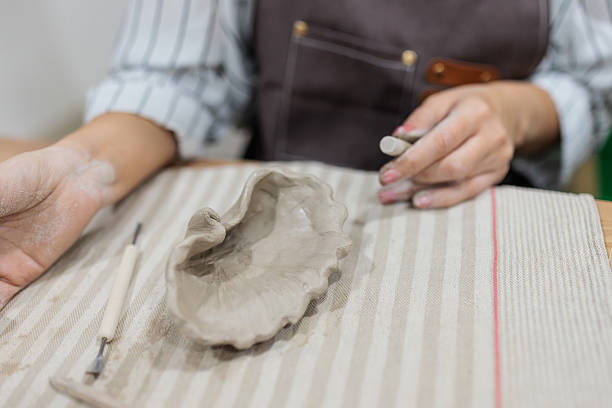 Detailed view of a shell-shaped clay bowl with the hands of the female artist covered in clay, showcasing her craftsmanship in a pottery workshop.