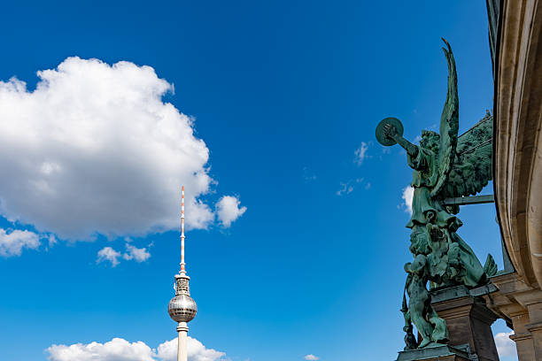 The Berlin TV Tower rises behind a striking angel statue. A bright blue sky with clouds forms the backdrop.