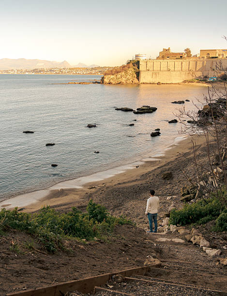 Back view of man standing on a coastal path with beach, sea, and historic walls in warm sunset light