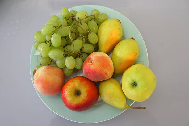 Green grapes (Vitis vinifera), a red apple (Malus domestica), yellow pears (Pyrus), and peaches (Prunus persica) are arranged on a light blue plate. The fruits exhibit varied colors: the grapes are a light green, the apple is red with touches of yellow, the pears are yellow with hints of red, and the peaches display a soft orangish-red hue. The background is a plain, neutral surface, highlighting the vivid colors and fresh appearance of the fruits.