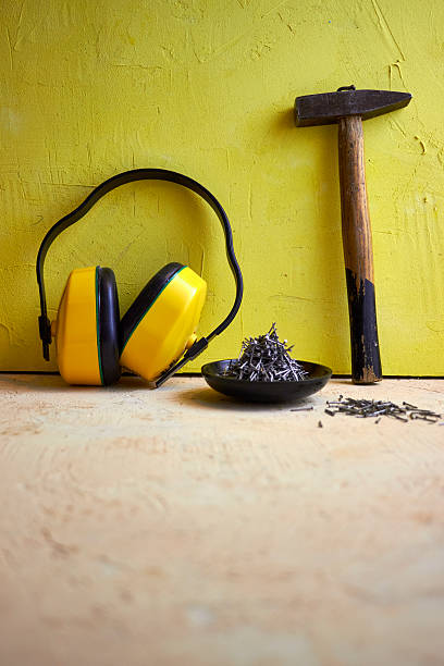 istock Still life with construction tools: hammer, nails in a wooden bowl, and protective earmuffs on a yellow textured wall background. Concept of building, DIY, safety, and repair work 2233397536