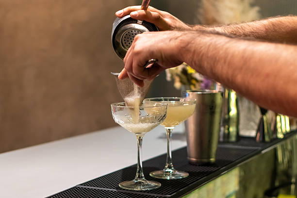 istock Bartender is expertly straining light-colored cocktail from shaker into two elegant coupe glasses. Other bar tools and blurred background are visible 2233411398