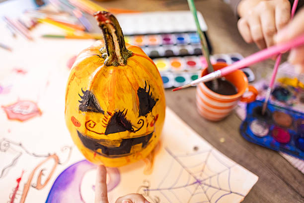 A close-up shot shows a cheerfully painted Halloween pumpkin, surrounded by art supplies and the female hands who are creating festive decor.