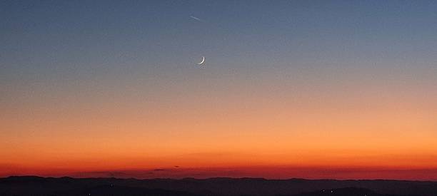 The crescent moon (silver of the moon) is visible during sunset with the enlightened trail of a plane, with a horizon made of dark mountains and the shades, or nuances, of blue, light blue, orange, and red colors