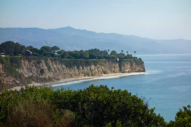 A Los Angeles Country Sheriff  helicopter flies over the dramatic, rugged cliffs of the Point Dume coast, where luxury homes are perched high above a secluded beach and the vast, blue Pacific Ocean.