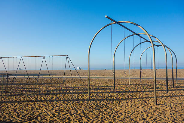 An empty and serene Santa Monica beach is captured on a clear morning, with a swing set and gymnastic rings in the foreground and a calm Pacific Ocean meeting the horizon.