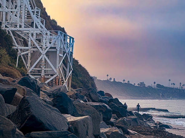 Encinitas, California, USA – 09/01/2025: A lone surfer carries a surfboard while walking near the iconic white steps leading to the Self-Realization Fellowship Center at Swami’s Beach during sunrise. The image highlights the coastal scenery and surf culture of Southern California.