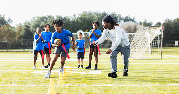 A black man coaching a co-ed flag football team, doing a practice drill on the field. He is watching and encouraging a 9 year old multiracial boy who is running with the football through a line of cones, while the other players watch.