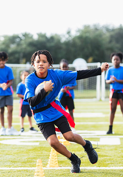 An Hispanic 10 year old boy on a co-ed flag football team at practice, running with the ball, zig-zagging through cones as his teammates watch.