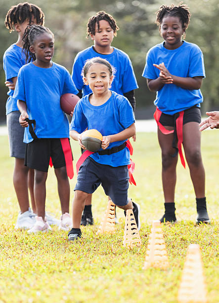 A multiracial 6 year old boy on a co-ed flag football team at practice, running with the ball, zig-zagging through cones as his teammates watch.