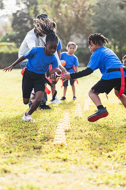 A coach with players on a co-ed flag football team at practice. A black 8 year old girl running with the ball, zig-zagging through cones as one of her teammates, a 10 year old Hispanic boy, tries to block her.
