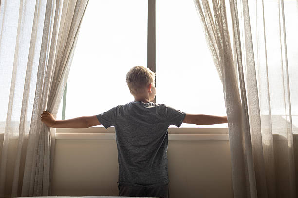 istock Boy Standing at Window Spreading Curtains to View Bright Outdoor Scene 2233442031