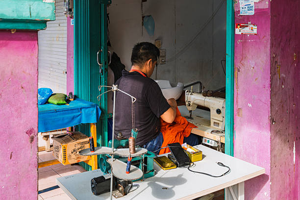 Balikpapan, Indonesia - August 3 2025. A candid shot of a male tailor diligently working at his sewing machine inside a small, colorful shop in Balikpapan, East Kalimantan.