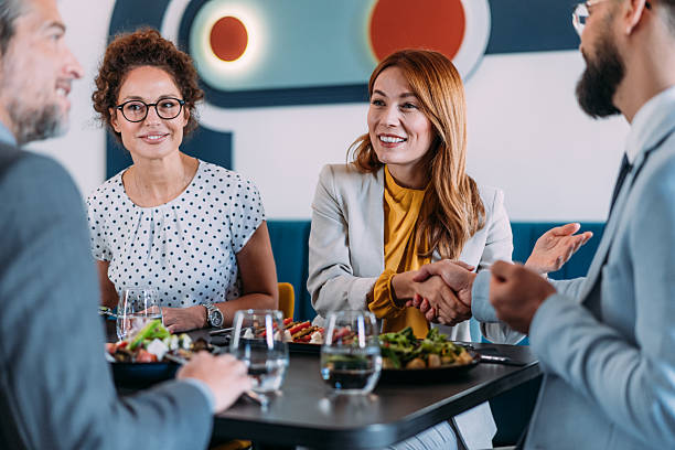 Shot of cheerful business persons shaking hands during business lunch or dinner at modern restaurant. Businesswoman and businessman shaking hands while having a lunch with their colleagues in a restaurant.
