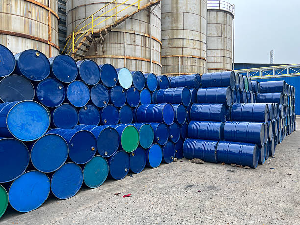 A pile of blue metal barrels stored outdoors at an industrial facility against a factory backdrop.