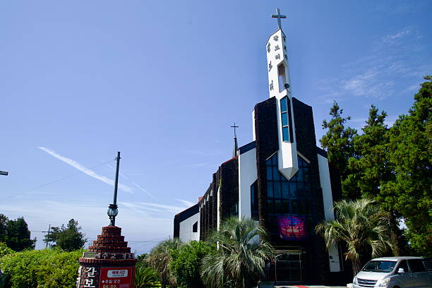 A modern church clad with dark basalt accents rises behind palms in Jungmun, its white steeple and crosses set against a clear sky along Jeju's southern coast.