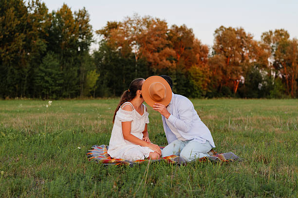 Two people share a sweet kiss while sitting on a blanket in an open field surrounded by trees at sunset