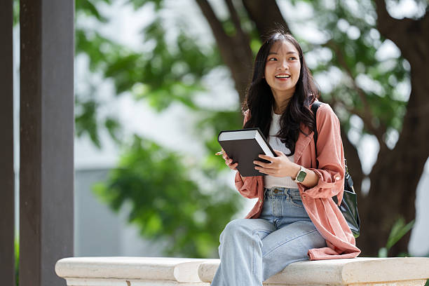 istock Happy young asian student sitting on a park bench, holding a book and smiling while enjoying the fresh air, immersed in study and preparing for upcoming exams 2233470803