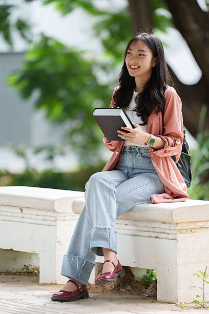 istock Happy young asian student is sitting on a bench, holding a book and smiling while looking away, enjoying the pleasant atmosphere of the campus 2233470807