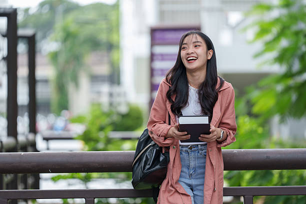 istock Cheerful young asian student is holding a book and smiling while leaning on a railing on a university campus, enjoying her educational journey 2233470841