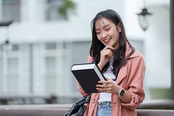 Young asian woman student smiling while reading a book outdoors on a university campus, embracing her studies and enjoying the vibrant atmosphere of academic life