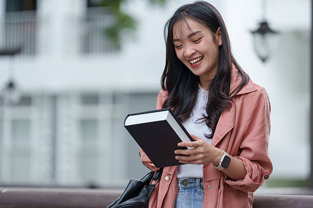 istock Cheerful young asian student is enjoying reading a book outdoors, laughing at something she has just read, with a backpack slung over her shoulder 2233470862