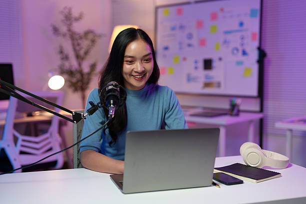 istock Smiling young asian woman recording a podcast in her home studio, using a professional microphone and laptop while surrounded by creative tools and illuminated by soft purple light 2233474255