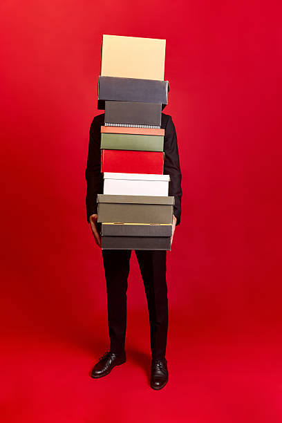 istock Person in black suit holding many boxes with hidden face on red background 2233474424