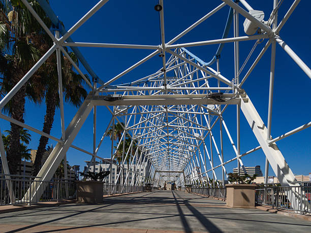 Intricate metal framework design of a pedestrian bridge under a vibrant blue sky surrounded by urban scenery and palm trees.