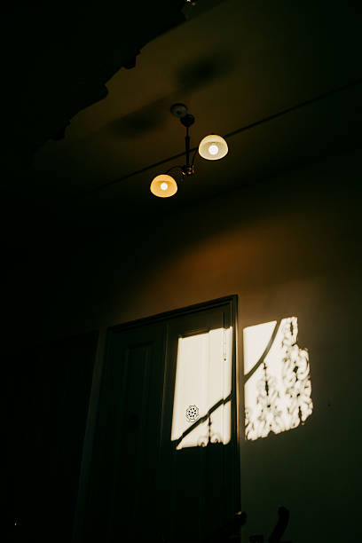 Sunlight casting decorative shadows on wall in dark room with ceiling lamp illuminating scene, creating mysterious and evocative atmosphere. Sunlight filtering through shadowy room, casting patterns