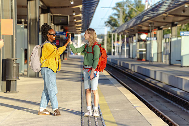 Two young women share a friendly high five while waiting at a sunny station platform.