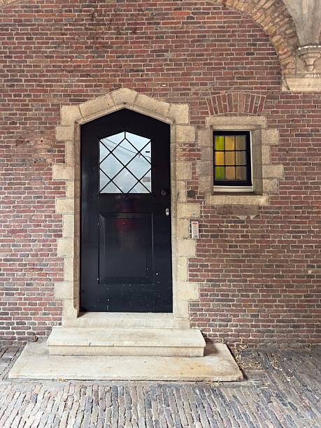 An arched door with a stone surround in a brick wall and a leaded window with a small stained glass window beside it.