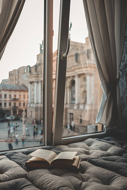 istock Cozy reading nook with an open book on a cushioned window seat overlooking a historic European city square and opera house at sunset through soft curtains 2233496754