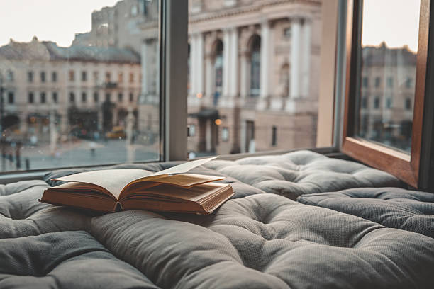 istock Cozy reading nook with an open book on a cushioned window seat overlooking a historic European city square and opera house at sunset through soft curtains 2233496756