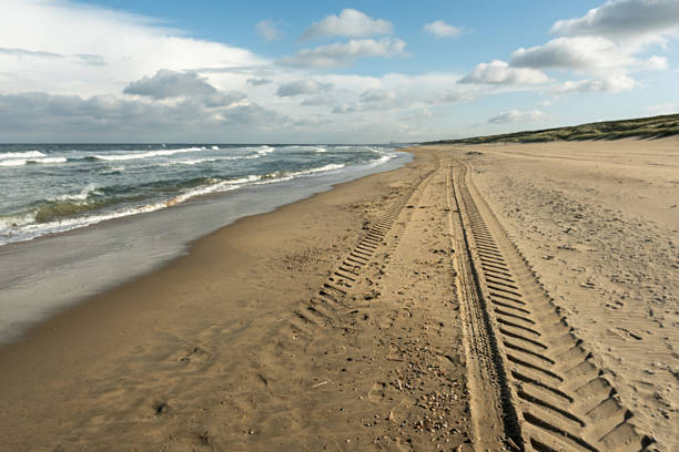 Deep tire tracks are imprinted on a sandy beach next to ocean waves on a partly cloudy day