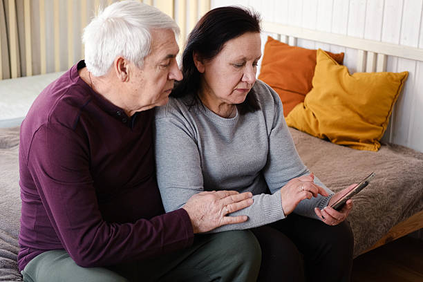 istock Old couple using a phone together while relaxing in the kitchen at home. Elderly man and woman spending leisure time browsing online with a smartphone. Happy senior married couple typing, learning 2233504571