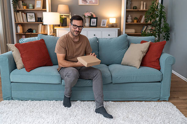 istock Man sitting on sofa holding a cardboard box and smiling at camera 2233511060
