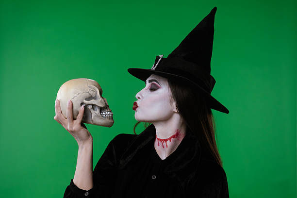 istock A vampire witch in a black cloak and hat poses with a skull on a green chroma key studio background 2233520810