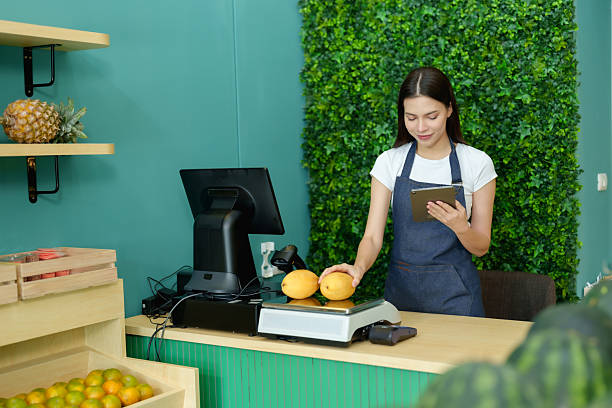 istock Caucasian female small business vendor checking fruit inventory using tablet while organizing counter with ripe mangoes on scale before opening retail store in modern grocery shop environment 2233522647