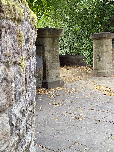 istock Stone gate pillars and a moss-covered curved wall lines a quiet, leafy path beneath a dense green canopy. Fallen leaves scatter the cobbled pavement, creating a peaceful, secluded atmosphere. 2233524608
