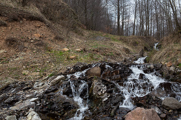 A cascading waterfall flows over dark rocks, surrounded by a hillside and bare trees, creating a scenic landscape.